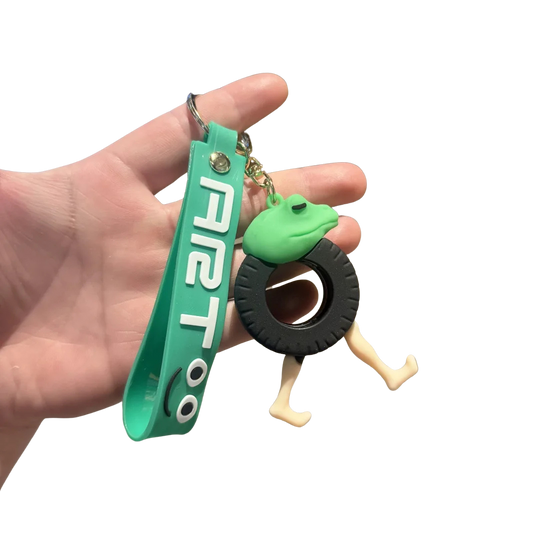 Hand holding a green keychain with a frog design and 'AIRT' branding on a white background